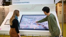 Zwei Personen stehen vor einem Bildschirm, der einen digitalen Stadtplan zeigt. Die Person rechts im Bild bedient den Bildschirm mit der Hand.