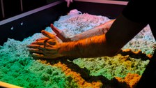Zwei Hände formen Sand in einer Box, darüber bunte Farben projiziert