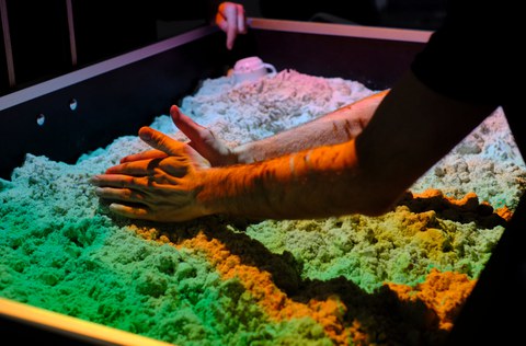 Zwei Hände formen Sand in einer Box, darüber bunte Farben projiziert