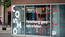Außenaufnahme des COSMO Wissenschaftsforums im Kulturpalast. Auf den Scheiben das COSMO-Logo und der Schriftzug "Forschung ganz nah erleben".