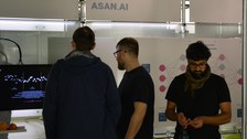 2 Personen stehen vor einem interaktiven Display, das die Software AsanAI vorstellt; eine weitere Person steht rechts davon frontal im Vordergrund. 