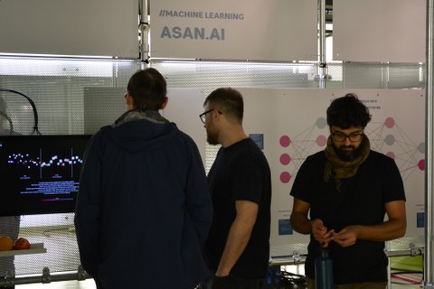 2 Personen stehen vor einem interaktiven Display, das die Software AsanAI vorstellt; eine weitere Person steht rechts davon frontal im Vordergrund. 