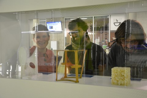 Foto von drei Gesichtern in Betrachtung der Ausstellungsexponate (im Vordergrund) durch die Vitrine hindurch fotografiert. 