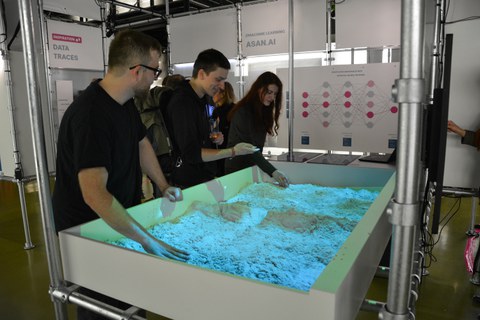 Drei Besuchende der Ausstellung entdecken die Funktionsweise von Schwarmverhalten am Exponat der interaktiven Sandbox.