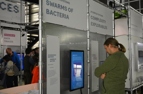 links: Blick in die Ausstellung mit mehreren Personen im Hintergrund; rechts: Eine Person beim Betrachten des Displays zum Thema Swarms of Bacteria  
