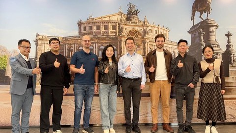 Photo: Michael Färber and his team stand together with employees of Xenon Automation Technology in front of a mural of the Semperoper opera house at the Xenon Automation Technology premises in China. They are giving a thumbs-up sign.