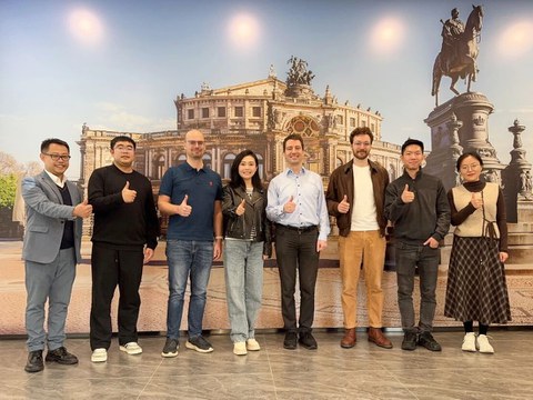 Foto. Michael Färber und sein Team stehen gemeinsam mit Mitarbeiter:innen von Xenon Automation Technology vor einem Wandbild der Semperoper in den Räumlichkeiten von Xenon Automation Technology in China. Sie zeigen einen Daumen nach oben.