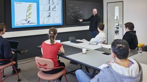 In a modern seminar room, a lecturer is giving a scientific presentation to a group of five participants. He stands in front of a blackboard with a sketched mathematical equation, pointing at a formula. Next to him, a large screen displays a graphical representation of genomic and geographic data. The audience, including a woman in a red top and four men in casual clothing, is seated at a table with laptops, notebooks, and coffee cups. The room is bright and functionally designed, with a black wall providing contrast.