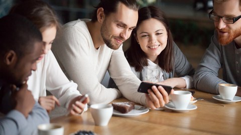 Five young adults are sitting around a table with coffee and cake, looking at smartphones