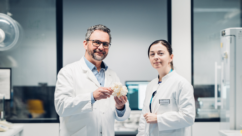 A photo. Two people wearing white labcoats pose for a picture in a laboratory-like setting. The person on the left holds a white grainy structure, the size of a handball ball, in his hands.