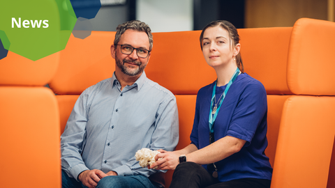 A photo. Two people sit next to each other on a futuristic orange sofa. The person on the right holds a white irregular shape.