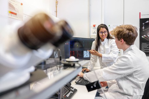 A photo. Two people are at the table with keyboard and screens. One person is sitting and his hands are laying on the keyboard. The other person stands next to him, talking, and gesturing. In the foreground there are blurred microscope oculars.