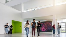 A photo. Seven people standing and walking in a large building hall. In the foreground 3 people talking to each other while walking. In the background: On the left, 2 people are engaged in a lively conversation, each holding a notebook in their hand. On the right, there are 2 people standing and talking, each carrying a backpack.