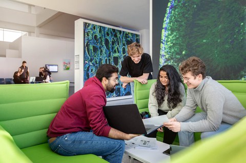 A photo. On the left side of the image, in the background, 4 people can be seen blurred. In the foreground, 4 people are chatting cheerfully sitting on green sofas that face each other. One person holds a laptop in his hand and presents its contents to the three people in front of him. One person stands leaning against the armrests of a sofa. Two people are sitting side by side on the sofa. One of them holds a sheet of paper in his hand. There are papers scattered on a table between the sofas.