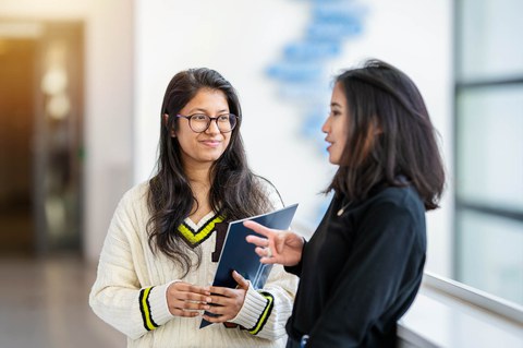 A photo. Two people standing next to each other. The person on the left is in focus. The person is facing the camera with their body and face, while the person on the right is standing sideways. The person in focus is holding a folder with the TU Dresden logo in their left hand and is looking at the person on the right with a friendly, attentive expression. The person on the right is speaking and gesturing.