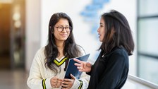 A photo. Two people standing next to each other. The person on the left is in focus. The person is facing the camera with their body and face, while the person on the right is standing sideways. The person in focus is holding a folder with the TU Dresden logo in their left hand and is looking at the person on the right with a friendly, attentive expression. The person on the right is speaking and gesturing.