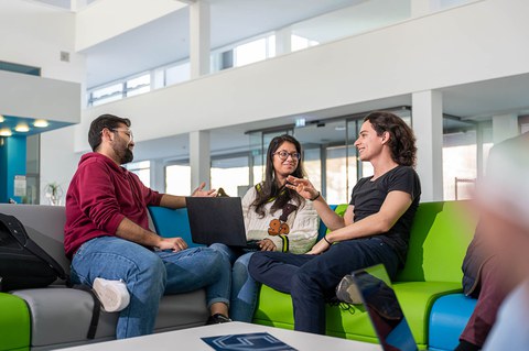 A photo. Three people are sitting on a corner sofa, talking with each other. The person in the middle has a laptop resting on their lap. The person on the left, as well as the person in the middle, are looking towards the person on the right. Everybody has cheerful faces and is engaged in lively conversation.
