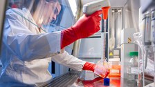 A photo. A person working in a laboratory. His hands are inside a glass cabinet. In one hand he holds a transparent square bottle with orange liquid. In the other hand, he holds an electronic pipette with long glass tip. The person has a concentrated facial expression.