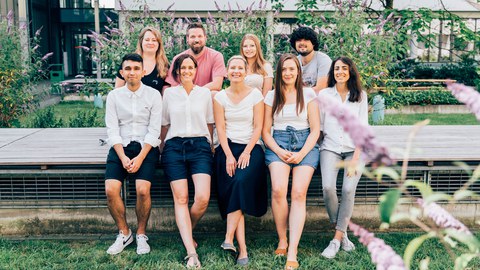 Group picture of 9 people sitting on a wooden bench in two rows. The scene is set in a green area with purple blooming bushes in the background.