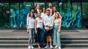 Group picture of 9 people standing in three rows on stairs in front of glass doors with a blue and green print on them.