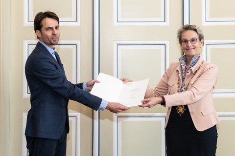 Zwei Personen. Ein Mann auf der linken Seite, der einen Anzug trägt. Eine Frau auf der rechten Seite, die ein formelles Outfit trägt. Gemeinsam halten sie eine Mappe in der Hand.