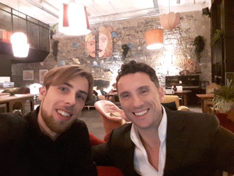 Two people sitting next to each other, smiling, in a rustic restaurant. They wear white shirts and black suits. In the background there are windows, wooden chairs and tables, and the room is lit by ceiling lamps. The walls are covered with wallpaper, which have a stone texture.