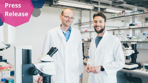 A photo. In the background, there is a bright laboratory with white walls and ceiling lights. In the center, two men wearing white lab coats are standing side by side and smiling at the camera. Behind them, there are laboratory benches with microscopes and various lab supplies, such as bottles, pipette boxes, and test tubes.