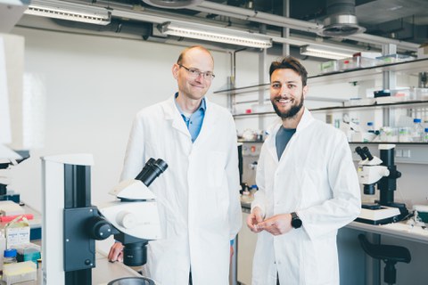A photo. In the background, there is a bright laboratory with white walls and ceiling lights. In the center, two men wearing white lab coats are standing side by side and smiling at the camera. Behind them, there are laboratory benches with microscopes and various lab supplies, such as bottles, pipette boxes, and test tubes.
