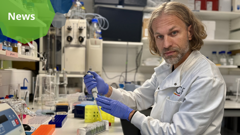 A photo. A male researcher in a white lab coat and violet gloves sits at a lab bench. He holds a pipette in his hands and looks directly at the camera. On the bench in front of him are lab equipment and a rack of small test tubes. 