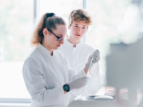 Ein Foto. Zwei Personen in Kitteln. Eine Person arbeitet mit einer Pipette, ungefähr auf Brusthöhe. Die zweite Person schaut gespannt über die linke Schulter zu. 