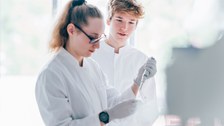 Ein Foto. Zwei Personen in Kitteln. Eine Person arbeitet mit einer Pipette, ungefähr auf Brusthöhe. Die zweite Person schaut gespannt über die linke Schulter zu. 