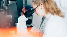 Ein Foto. Zwei Personen vor einem Mikroskop. Eine schaut durch das Okular, die andere steht daneben. Im Vorderung ist ein unscharfes organges Objekt, welches den unteren linken Teil des Bildes verdeckt. Im Hintergrund sind weitere Laborgeräte zu erkennen.
