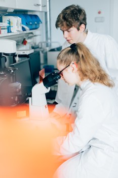 Ein Foto. Zwei Personen vor einem Mikroskop. Eine schaut durch das Okular, die andere steht daneben. Im Vorderung ist ein unscharfes organges Objekt, welches den unteren linken Teil des Bildes verdeckt. Im Hintergrund sind weitere Laborgeräte zu erkennen.