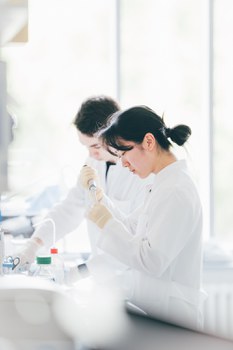Ein Foto. Zwei Personen in Kitteln. Die Person im Vordergrund abeitet mit einer Pipette, ungefähr auf Brusthöhe. Die Person dahinter arbeitet auf einem Tisch der vor beiden Presonen steht. Beide Personen haben ein konzentriertes Gesicht.