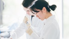 Ein Foto. Zwei Personen in Kitteln. Die Person im Vordergrund abeitet mit einer Pipette, ungefähr auf Brusthöhe. Die Person dahinter arbeitet auf einem Tisch der vor beiden Presonen steht. Beide Personen haben ein konzentriertes Gesicht.