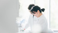 Ein Foto. Zwei Personen in Kitteln. Sie stehen an einem Tisch und vor Ihnen hängt ein Schrank. Dieser Tisch und Schrank befinden sich in der linken Bildhälfte und sind unscharf. Auf dem Tisch stehen verschiedene Laboruntensielien. Mit diesen arbeiten die beiden Personen.