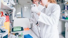 Ein Foto. Zwei Personen in Kitteln. Eine Person arbeitet mit einer Pipette, ungefähr auf Brusthöhe. Die zweite Person schaut gespannt über die rechte Schulter zu. Vor den zwei Personen steht ein Tisch mit vielen Forschungsutensilien, zudem hängt ein Schrank über dem Tisch mit weitern Forschungsutensilien.