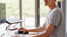 Ein Foto. Eine Person steht an einem Schreibtisch und hat die Hände an Tastatur und Maus. Vor der Person ist ein Bildschirm zu sehen.