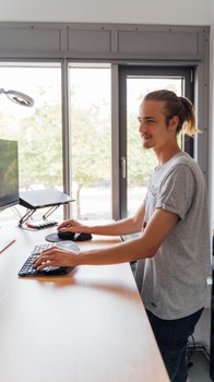 Ein Foto. Eine Person steht an einem Schreibtisch und hat die Hände an Tastatur und Maus. Vor der Person ist ein Bildschirm zu sehen.