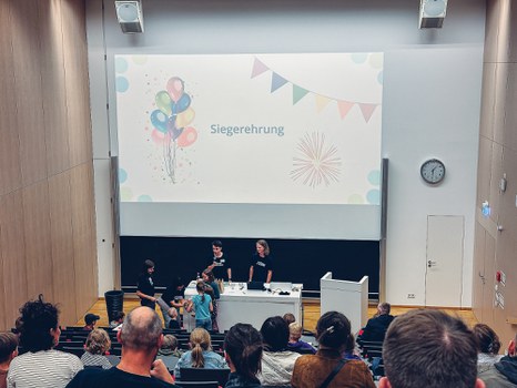 Ein Foto. Ein Hörsaal mit einigen Menschen. Über der Tafel ist der Schriftzug „Siegerehrung“ zu sehen. Vor der Tafel, am Rednerpult bekommen zwei Kinder Geschenke.