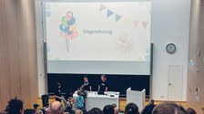 Ein Foto. Ein Hörsaal mit einigen Menschen. Über der Tafel ist der Schriftzug „Siegerehrung“ zu sehen. Vor der Tafel, am Rednerpult bekommen zwei Kinder Geschenke.
