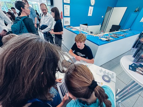 Ein Foto. Im Fokus sind 3 Personen. Ein Person sitzt an einem Tisch und spricht mit den zwei anderen Personen. Diese sind nur von hinten zu sehen, man sieht nur deren Köpfe. Die zwei Personen halten einen Zettel in der Hand. Im Hintergrund unterhalten sich Personen.