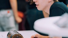 Ein Foto. Eine Person, welche etwas ausserhalb des Bildes anschaut. Die Person sitzt an einem Tisch, vor ihr steht eine Schatztruhe, Stifte, Zettel und Flyer.