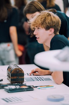Ein Foto. Eine Person, welche etwas ausserhalb des Bildes anschaut. Die Person sitzt an einem Tisch, vor ihr steht eine Schatztruhe, Stifte, Zettel und Flyer.