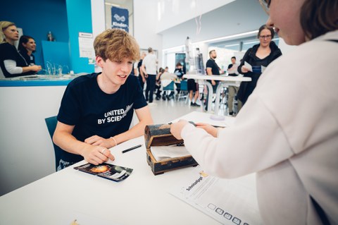 Ein Foto. Zwei Peronen sind zu sehen. Eine Person sitz am Tisch, die andere steht vor dem Tisch und öffnet eine auf dem Tisch stehende Schatztruhe. Das Bild wurde über die linke Schulter der stehenden Person gemacht, somit ist die stehende Person weniger sichtbar.