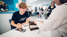 Ein Foto. Zwei Peronen sind zu sehen. Eine Person sitz am Tisch, die andere steht vor dem Tisch und öffnet eine auf dem Tisch stehende Schatztruhe. Das Bild wurde über die linke Schulter der stehenden Person gemacht, somit ist die stehende Person weniger sichtbar.