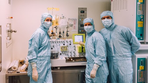 Drei Personen mit Schutzanzügen, Handschuhen, Masken und Hüten posieren vor einer Maschine in einer Laborumgebung. An der Maschine sind Infusionsbeutel angeschlossen.