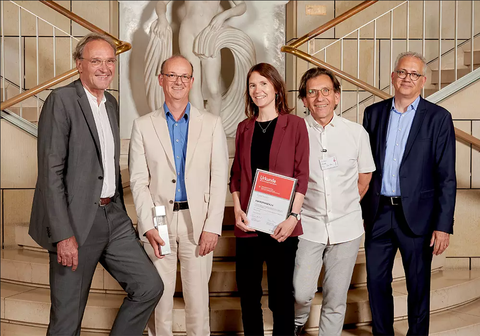 Ein Gruppenfoto mit fünf Personen. Die beiden Personen in der Mitte halten ein Diplom und einen Preis, der wie ein Block aus Metall und Glas aussieht.