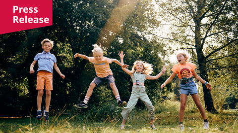 A photo. Four children are captured mid-jump in an outdoor setting with a background of dense green trees and sunlit grass in the foreground. They are wearing colorful summer clothing including striped and solid orange shirts, shorts, and a patterned jumpsuit. Each child is shown in a dynamic jumping pose with arms and legs outstretched. A bright sunbeam cuts diagonally across the center of the frame from the top. In the top left corner, a magenta rectangular banner contains the white text "Press Release"