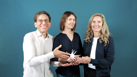 A photo. Portrait of three people smiling. They hold a white shape, looking like made of porcelain, together in their hands. 